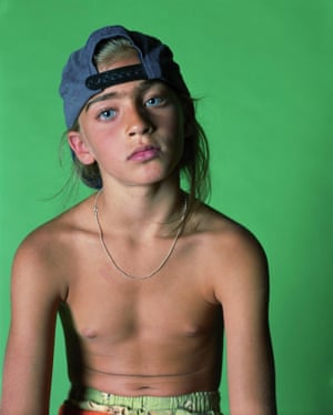A girl aged about seven or eight with no shirt on and wearing a backwards baseball cap and shorts and looking at camera with a green backdrop