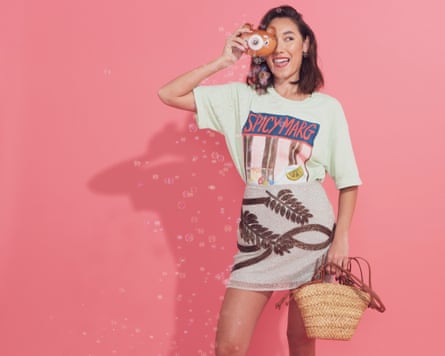 A woman wearing a T-shirt and beaded skirt and holding a woven straw bag holds a bubble camera up to her eye. She is surrounded by bubbles and is shot against a pink background