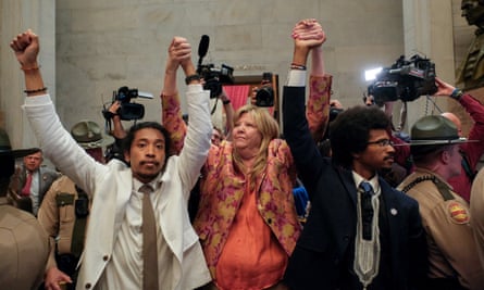 From left, a young Black man with long black hair pulled back and wearing a white suit, a middle-aged white woman with blond hair wearing a peach outfit, and a young Black man in glasses wearing a dark suit, raise their linked hands. They are in a large, white marble room surrounded by state troopers and people holding video cameras.
