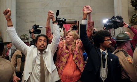 From left, a young Black man with long Black hair pulled back and wearing a white suit, a middle-aged white woman with blond hair wearing a peach outfit, and a young Black man with an Afro and glasses wearing a dark suit, raise their linked hands. They are in a large, white marble room surrounded by state troopers and people holding video cameras.