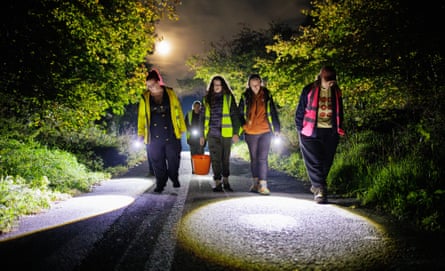 Four women in hi-vis vests, carrying torches and a bucket, walk down a dark country lane flanked by shrubs and trees