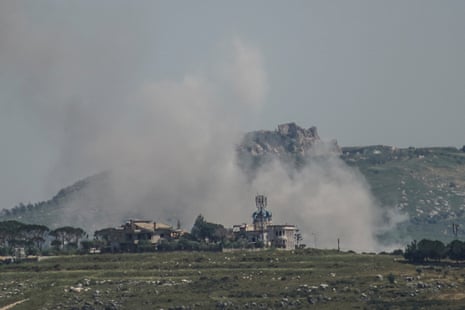 Smoke rises from behind some buildings against a backdrop of a large hill and grey sky.