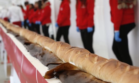 A huge baguette is displayed on a long table with people in the background