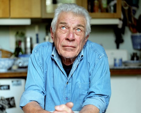 Writer John Berger leans on a table in a kitchen, hands clasped, looking upward