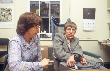 Stephen Hawking in his younger years, sits in a wheelchair in a room alongside an unidentified woman