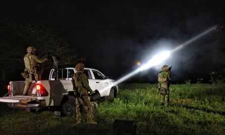 Night-time picture of a soldier in uniform aiming a heavy machine gun mounted on the back of a white civilian pickup truck, with other soldiers around him