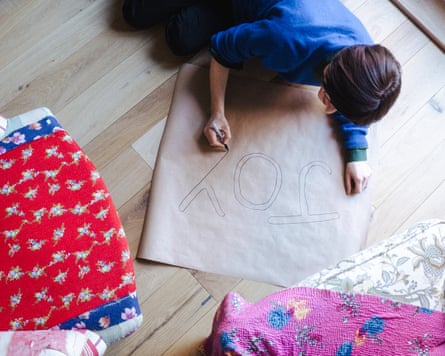 Emma liegt auf dem Boden und zeichnet mit einem Filzstift die Buchstaben JOY auf ein großes Blatt Papier.