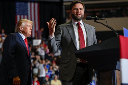 A man wearing a gray suit speaks at a podium with his hand gestures in the air, as a man in a navy suit looks on at the crowd