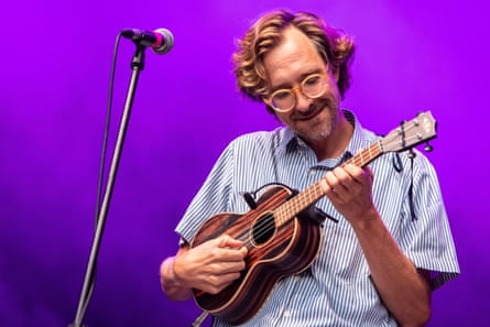 A man wears a blue shirt and plays a small guitar against a purple backdrop.