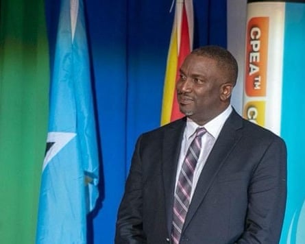 a formal photo in which he stands in front of flags, wearing a suit and tie. He has a close-shaven head and neat, short grey beard.