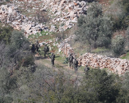A high wide shot of several Israeli soldiers in green fatigues on a path by a stone wall and hillside