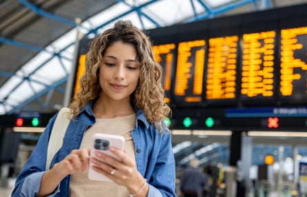 A young woman in a T-shirt and blue shirt, checking her phone at a train station