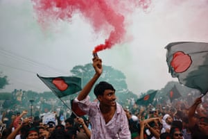 A man in the foreground holds up a red smoke flare as a crowd behind him wave Bangladeshi flags