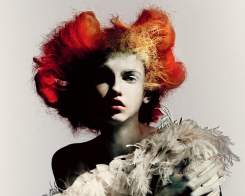 A model with voluminous red and orange hair looks upward, wearing a white feathered garment