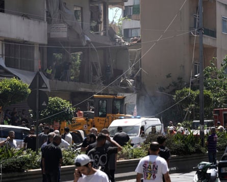 A crowd forms in a street around an ambulance parked outside a badly damaged building