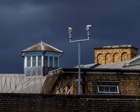 A view of rooftops and surveillance cameras at HMP Wandsworth in London, with a brick wall covered in coils of barbed wire in the foreground. The sky above is dark grey.