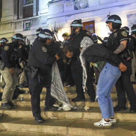 officers in helmets on steps hold on to people wearing keffiyehs. at least one appears to have their hands tied together, in a color photo