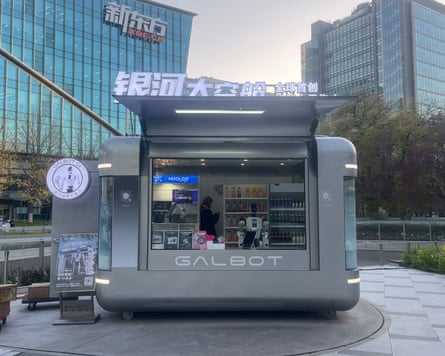 A silver vending booth with a humanoid robot serving. In the background is a human overseer.