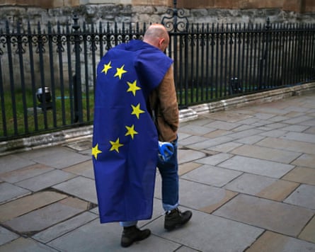 A man with the EU flag wrapped around him as a cape walks despondently along the pavement with his hands in his pockets.