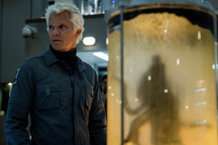 Timothy Olyphant standing in front of a glass cell containing an alien, which is silhouetted against the glass
