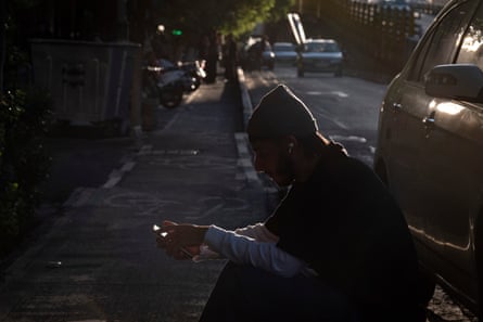 A young Iranian man uses his mobile phone while sitting on a pavement next to a car