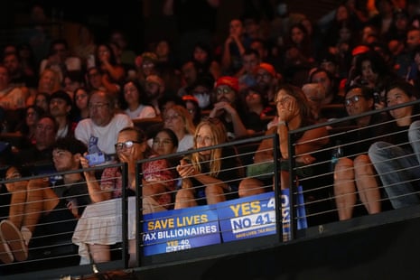 Rally crowd listens to Bernie Sanders, with "Save Lives, Tax Billionaires" signs on the railing