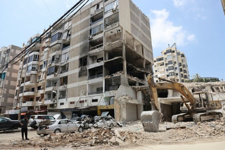 A man speaks on a mobile phone while standing near a destroyed apartment block. An excavator is parked alongside the building