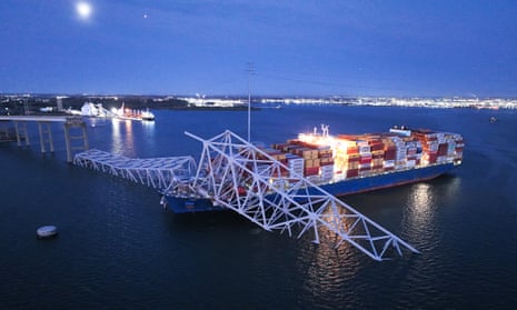 A collapse bridge partly rests on an enormous cargo ship in a bay with a city shining in the distance.