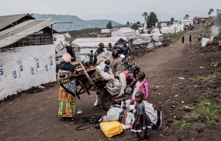 A man and woman, standing on a dirt road, load belongings on to a motorbike as two young children stand alongside. Makeshift tents are behind.