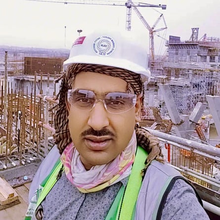 A selfie of a young South Asian man wearing a hard hat at a building site