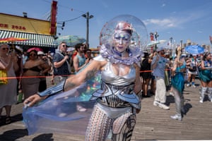 A woman wearing a mermaid costume and a transparent sphere around her head, with full face makeup, pink and silver dyed hair and tattoos, looks straight at the camera