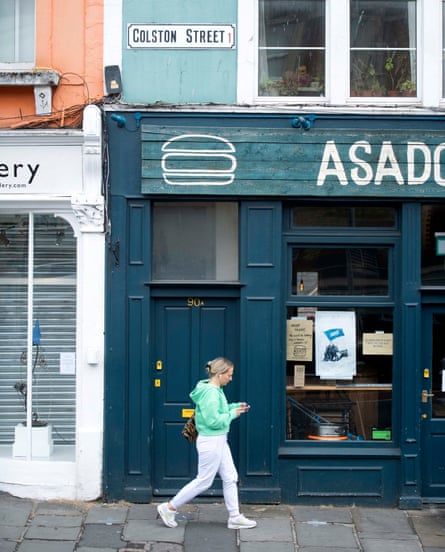White Colston Street sign on a turquoise painted section of wall above a cafe with dark teal-coloured woodwork, sign, doorway and windowframes. A woman is walking past looking at her phone.