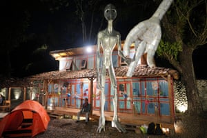 A man sits on the verandah of a building, outside which is an alien sculpture/design