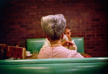 The back of a grey-haired woman wearing a pink dress and pearls. She is sitting at a diner table in a restaurant, on a green banquette, with a brick wall facing her