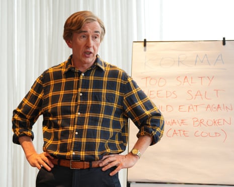 Steve Coogan in character as Alan Partridge. He stands in front of a flipchart, wearing a checked shirt, with this hands on his hips