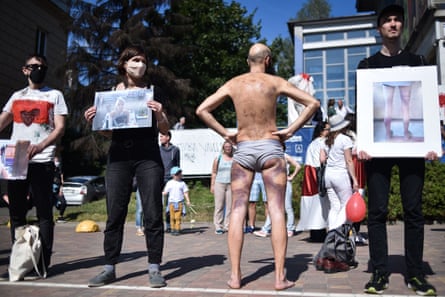 A man wearing only underwear stands with his back to the camera to display bruises on his legs and back, while three other men face the camera holding photos of beaten men