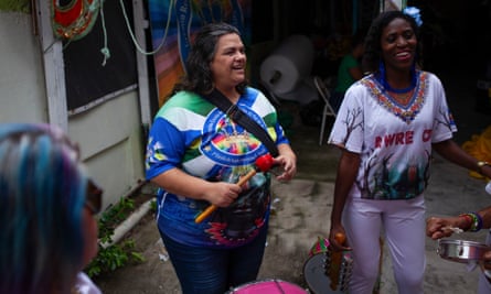 A women in jeans and a samba school top holds a drum stick and laughs with other women