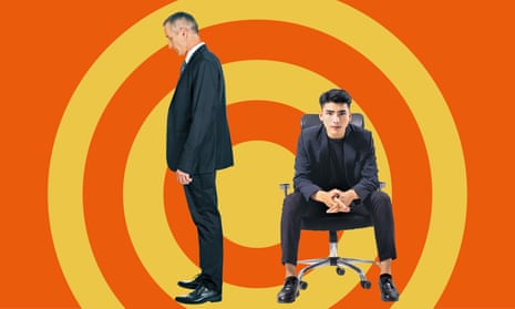 One man standing looking downwards, another man sitting on a chair