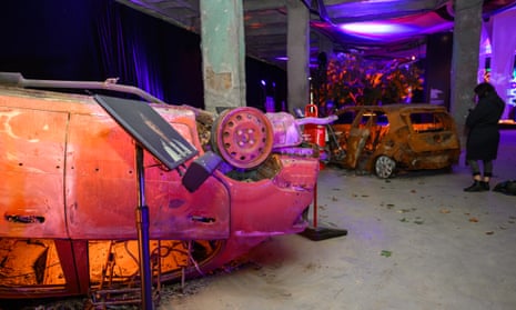 Two totally burnt out cars, one upside down, illuminatewd with orange light, form part of an exhibit