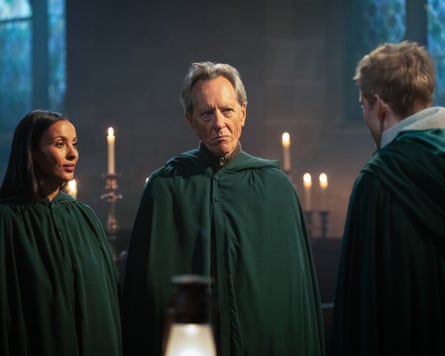 A woman and two men stand in a church lit by candles wearing green cloaks