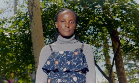 Vanessa Nakate, climate activist and author, poses for a portrait outside of the United Nations headquarters. Nakate is standing in front of some trees and wearing a grey turtleneck underneath a dark blue dress with a colourful print pattern on it