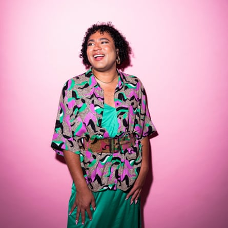 Travis Alabanza in a green dress and patterned blouse against a pink background