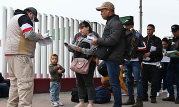 Under a cloudy sky, a line of adults and children wait in an orderly line on pavement as a man facing them fills out papers on a clipboard.
