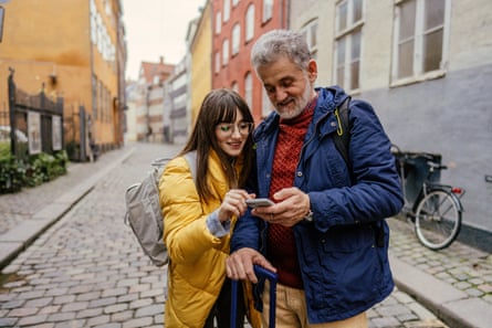 A young woman guides an older man as he holds a smartphone; she has long dark hair and wears a yellow coat while he has grey hair and beard, wears a blue anorak and leans on a stick. They are standing on a cobbled street in a historic part of a Danish city with colourful houses and bicycles in the background.