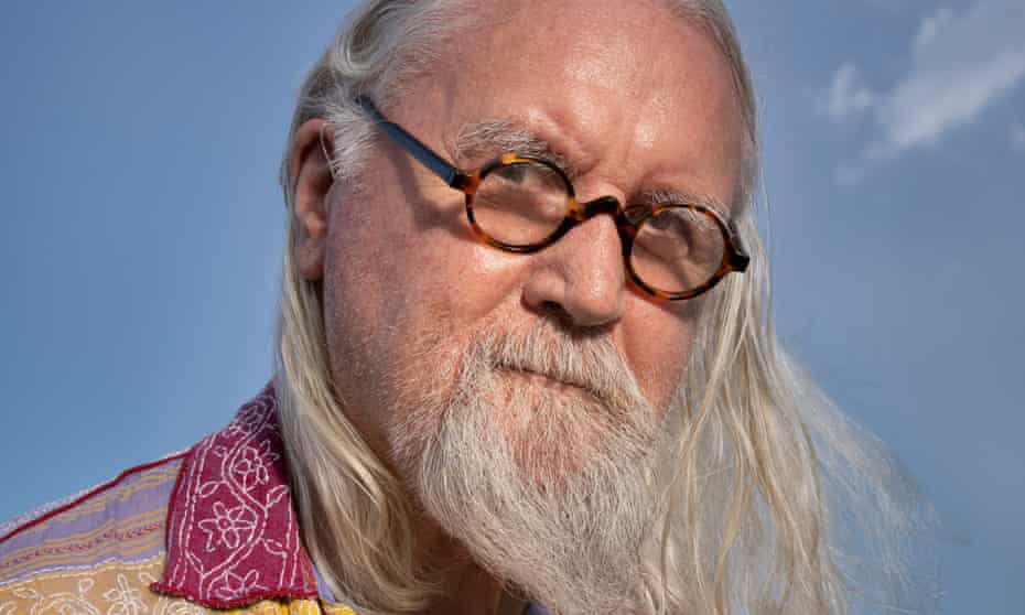 Billy Connolly, with long hair, long beard and glasses, looking into the camera, blue sky behind him