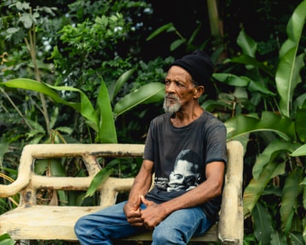 Williams sits on a wooden bench in front of lush, green vegetation; he is thin and has a grey beard; he wears a dark grey T-shirt with an image of Malcolm X on the front, a black woollen hat and blue jeans