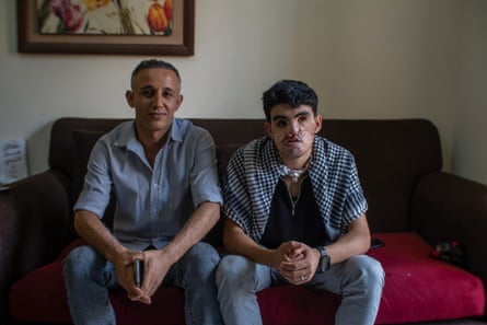 A man and a teenage boy with facial injuries sit side by side on a red sofa.