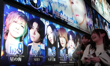 Two young women laughing as they pass by a brightly lit billboard of photos of enticing-looking young men at a host club in Tokyo