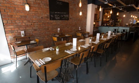 A long wooden table with metal-framed wooden chairs and, on the brick wall, a chalkboard with writing on it