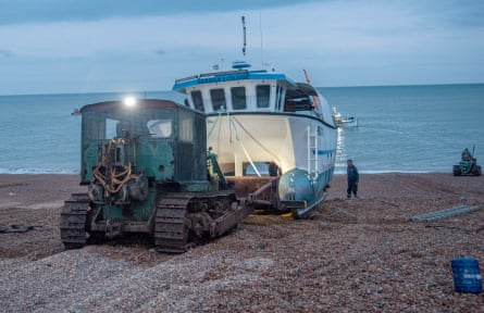 A fishing boat is moved on a shingle beach at dawn by a vehicle with caterpillar tracks